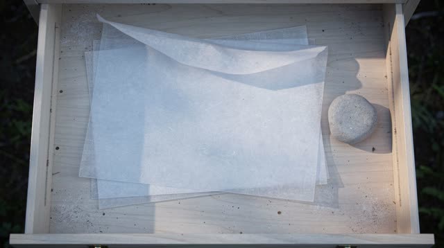 Airy top-down crop of a shallow park map drawer after rain; blank translucent vellum sheets rest on dry pale wood, bendi