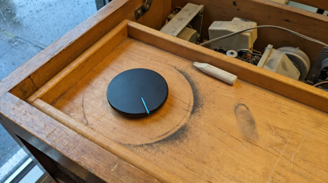 Tight oblique crop inside a public radio repair cabinet after rain; one blank matte black tuning dial rests in a shallow