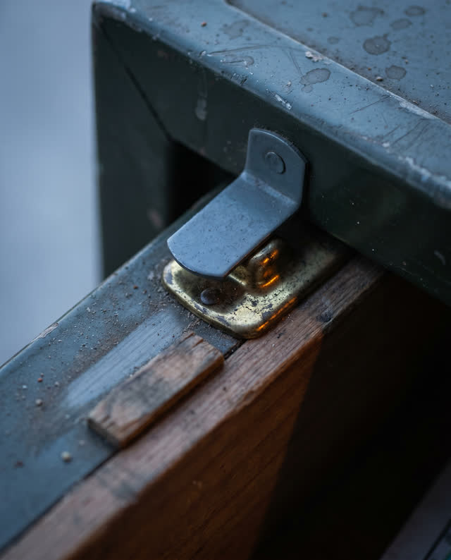 Tight asymmetric macro crop of an early blue-hour park map case repair drawer after rain; one dull slate-gray latch tab