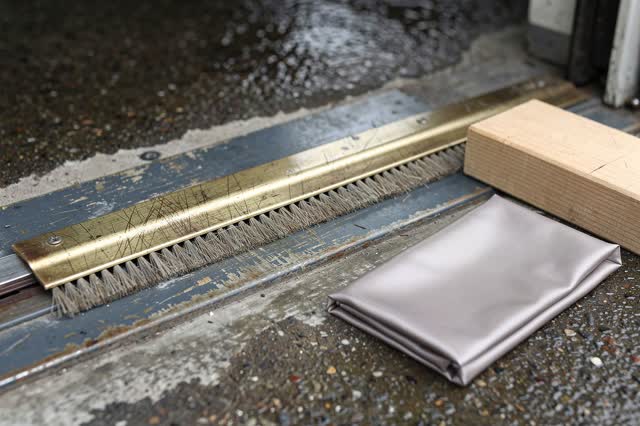 Low horizontal process crop at a covered transit-shelter doorway threshold after rain; one bent dull-brass door-brush st
