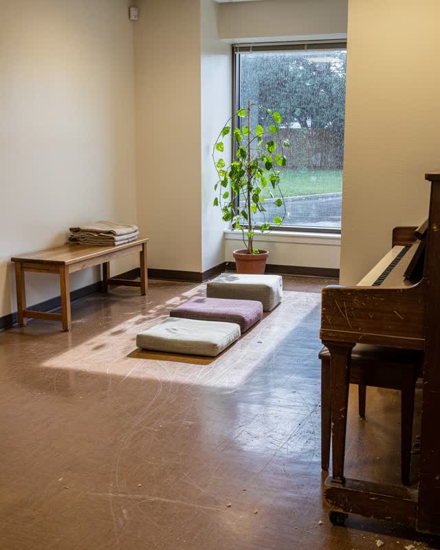 Medium-wide view of a small public learning commons after rain; a worn piano edge with one blank practice card, a civic