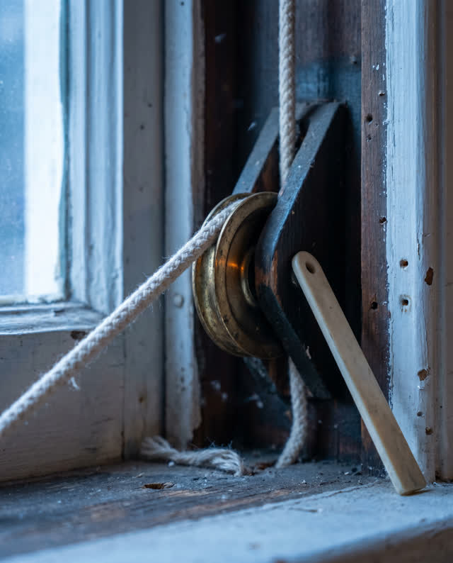 Tight asymmetric crop inside an old branch-library window-weight pocket at early blue hour; one small brass pulley wheel