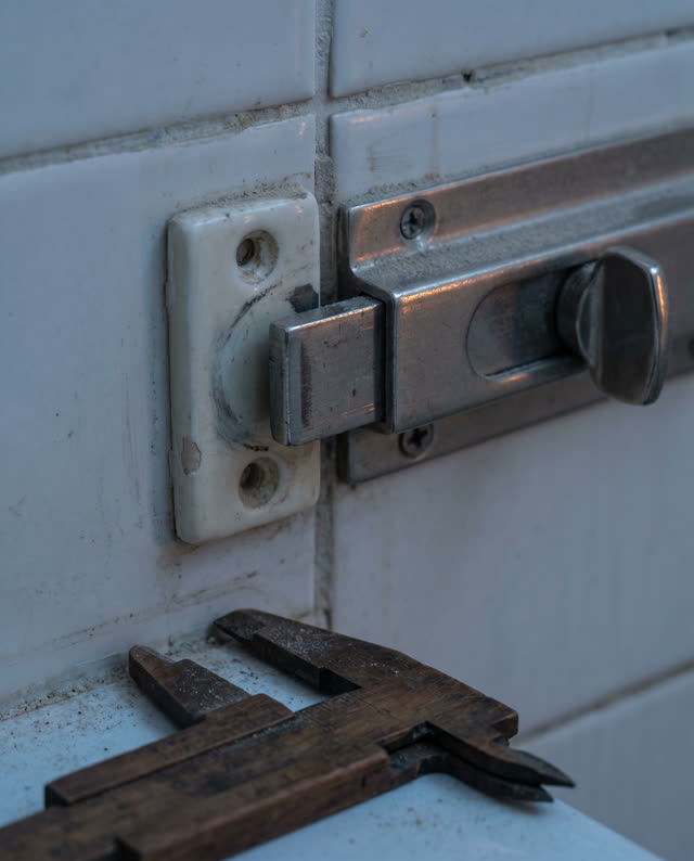 Tight asymmetric crop of an old public shower privacy-latch plate at early blue hour, dry after service; one dull nickel