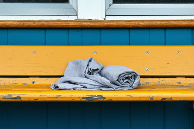 Restful low horizontal crop on a civic coatroom bench near a high frosted window after rain; one folded blank canvas cla