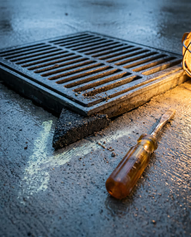 Tight low asymmetric crop on a small public utility floor drain after rain, with no room visible. One corner of a dark g