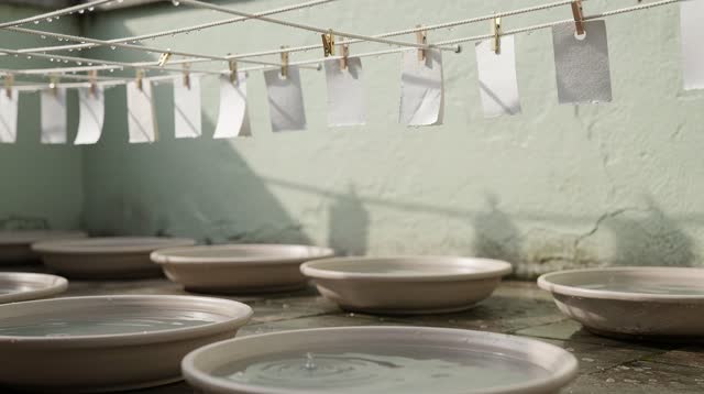 An open-air civic drying court after rain, pale walls and cotton lines strung overhead. Blank paper weather tags behave