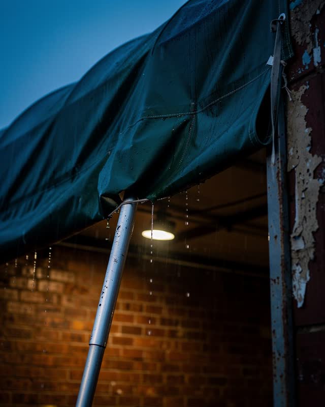 A civic loading-bay awning during steady rain at early blue hour, seen from an asymmetrical upward angle beneath the fab