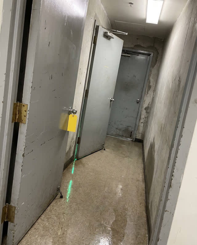 A narrow back corridor after rain with three heavy service doors failing to close in sequence. Misaligned door closers p