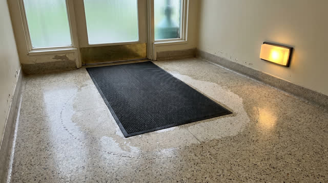 A small neighborhood clinic vestibule just after rain, before opening, viewed slightly above floor level with clear read