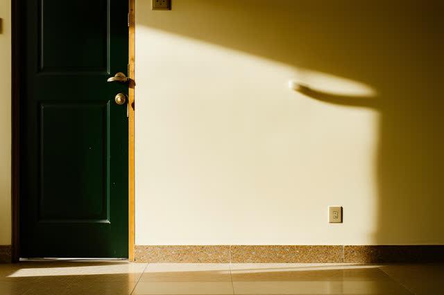 A plain community room doorway in thin late-afternoon light. One worn rubber doorstop holds a half-open painted door, ke