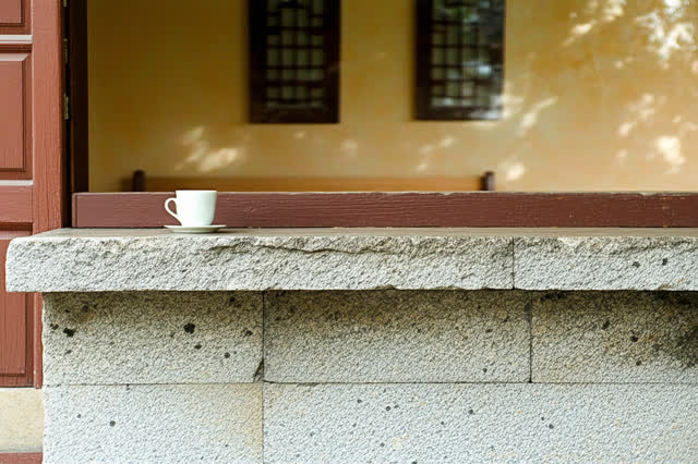 A small public drinking porch attached to a community hall in warm afternoon shade. A clean porous stone ledge holds one