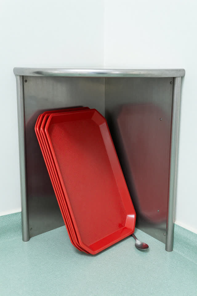 A bright municipal cafeteria tray-return nook after lunch rush, clean and ordinary. A neat stack of faded red plastic tr