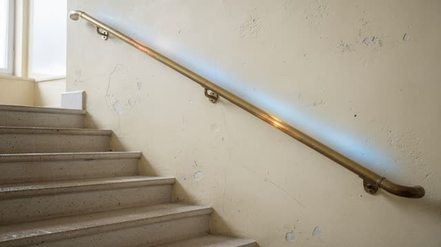 A sunlit public stairwell landing in an old civic building after morning rain. A dull brass handrail runs diagonally thr