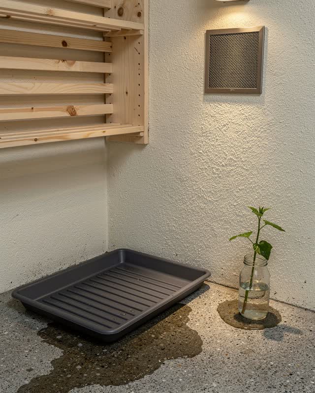 A neighborhood workshop mudroom after rain, designed for wet things to return gently. A shallow matte charcoal boot pan