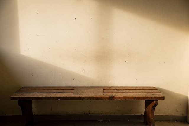A small public waiting alcove in gentle late-afternoon side light. A long worn wooden bench sits empty against a cream w