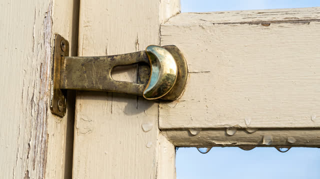 A bright side entrance to a shared workshop after rain, focused on a worn brass door latch and striker plate. The latch