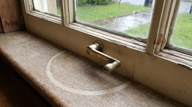 A bright public reading-room side window after rain. A dull brass window crank sits off-center on a cream-painted frame,