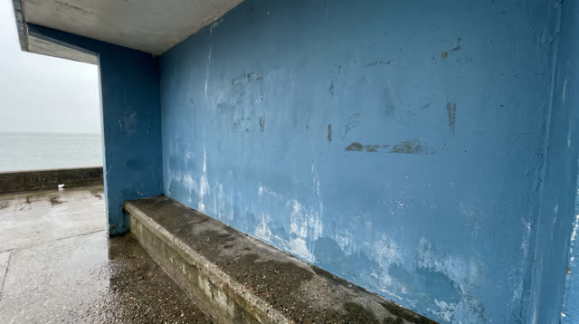 A small coastal ferry shelter after rain, open to gray waterlight but empty of people. A broad chalky blue painted wall