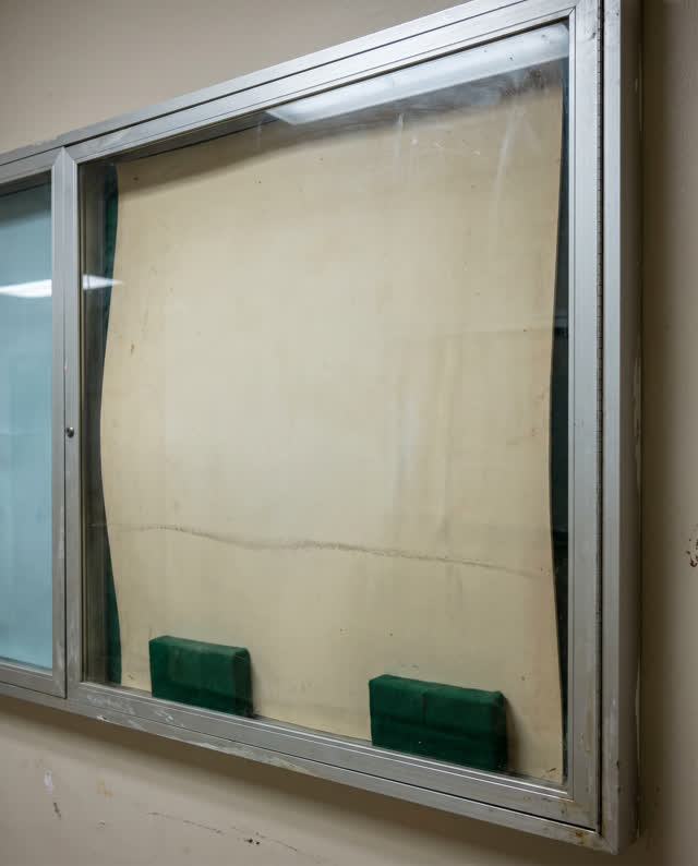 A public hallway display case with all contents removed and no readable notices. Inside the empty case, a thin dull crea