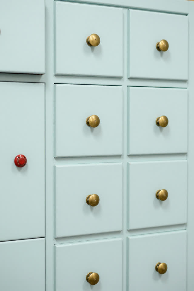 A bright public craft-room storage cabinet, clean and ordinary. Several dull brass drawer pulls have shifted by tiny dif