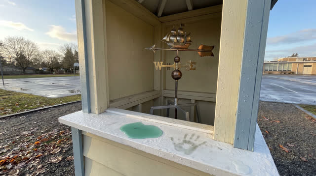 A small public weather kiosk on a schoolyard edge after rain, empty of people and with no readable signage. Inside the s