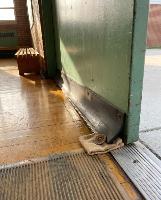A school gym side door after rain cleanup, empty of people, with no readable signs, threshold mat repetition, bench slat
