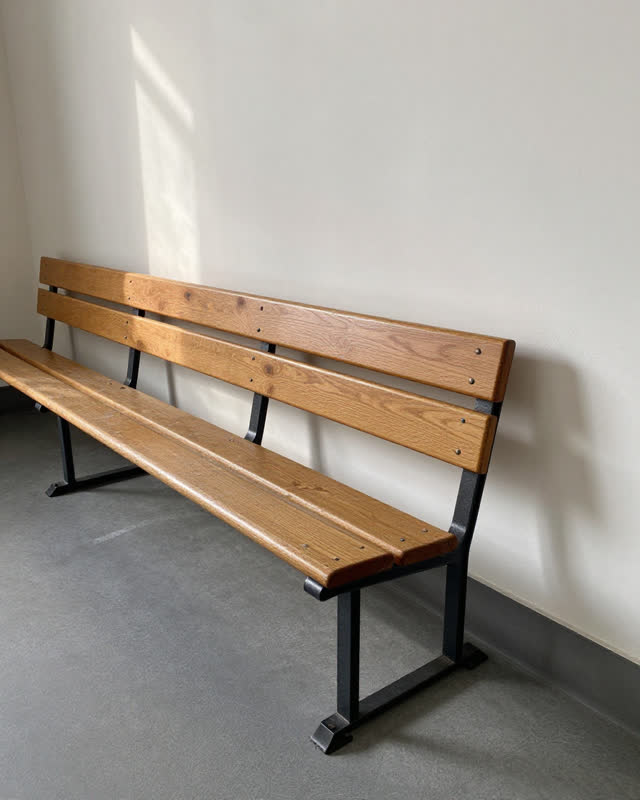 A public waiting-room bench row near a sunlit wall, empty of people, with no readable notices, library-cart repetition,