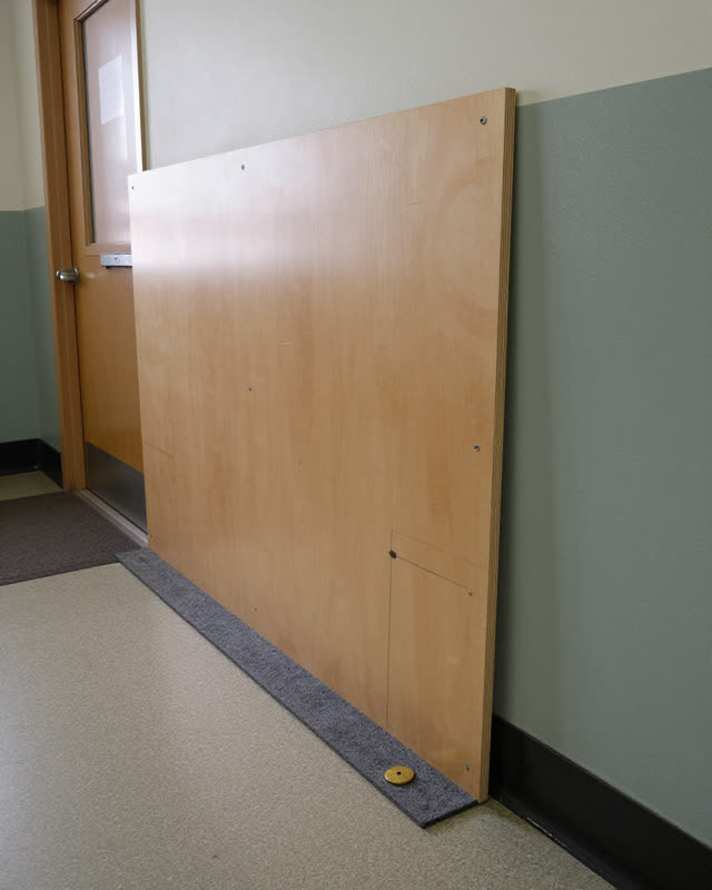 A school back hall with a temporary display backing panel leaning beside a service alcove, not a notice case, empty of p