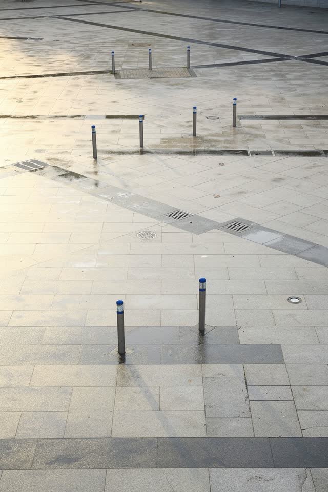 A bright playful open public plaza after morning rain: wet paving seams and the crisp shadows of movable painted-metal b