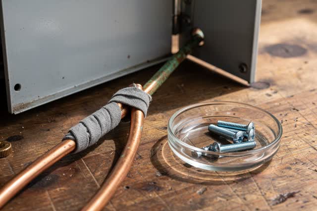 A tender practical repair scene on a small daylight workbench: an old copper cable is being sleeved with gray cloth and