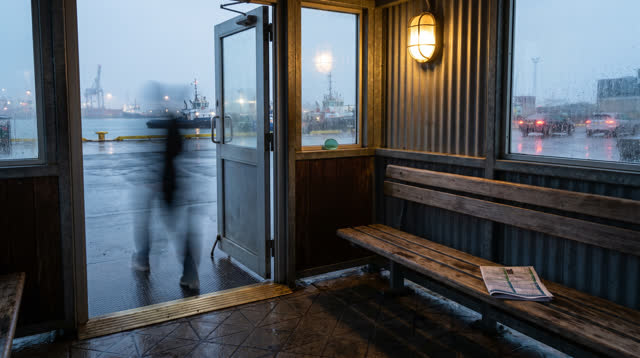 A lucid self-portrait as a rain-silver ferry shelter beside a working harbor, inhabited by traces of use but not crowded