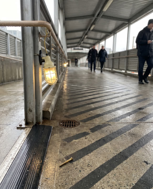 A tense covered public walkway after rain: louver shadows slip out of alignment across the wet floor, pulling diagonally
