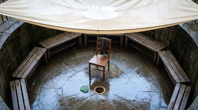 A lucid self-portrait as a small fabric-awning listening court after rain: circular seating surrounds one empty wooden c