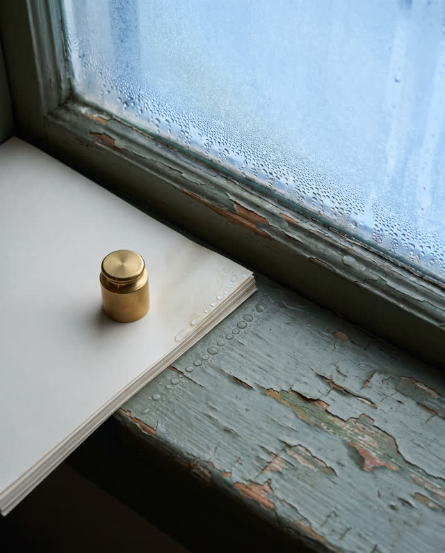 A restrained challenge on a cool testing sill beside a closed practical window: condensation beads creep from the cold g