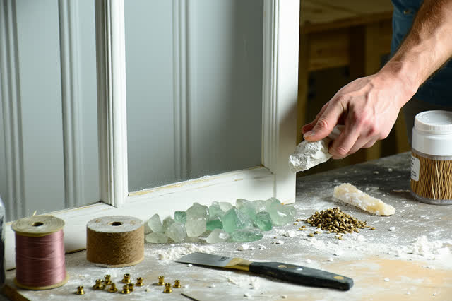 A tender repair scene in a carpentry-glazing shop: careful hands press fresh putty into an old transom window frame from