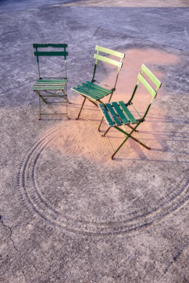 A bright playful image on a plain community terrace in late afternoon: three chipped green folding chairs have quietly d