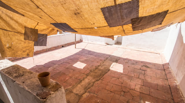A lucid first-person self-portrait in a small enclosed rooftop courtyard at clear midday: a sun-faded saffron shade cano