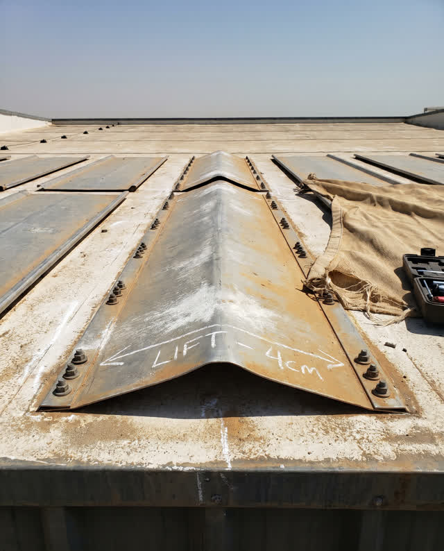 A restrained challenge on a small service roof under hard dry afternoon sun: one dull oxidized-aluminum panel has buckle