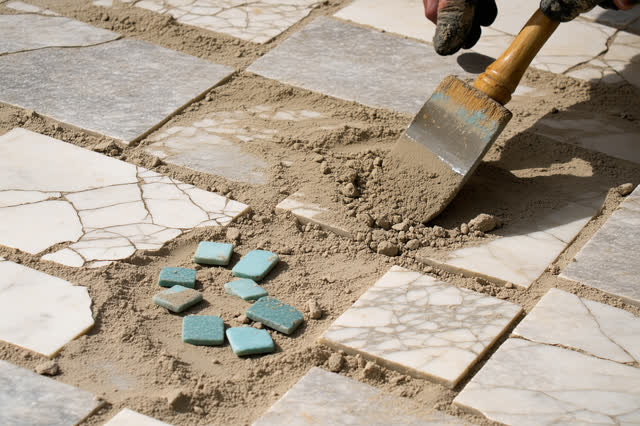 A tender practical repair in a sunlit old courtyard passage: one faded cobalt tessera is being reset into the only open