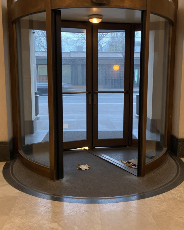A restrained challenge in a public entrance vestibule before dawn: one leaf of a three-part revolving door has drifted v