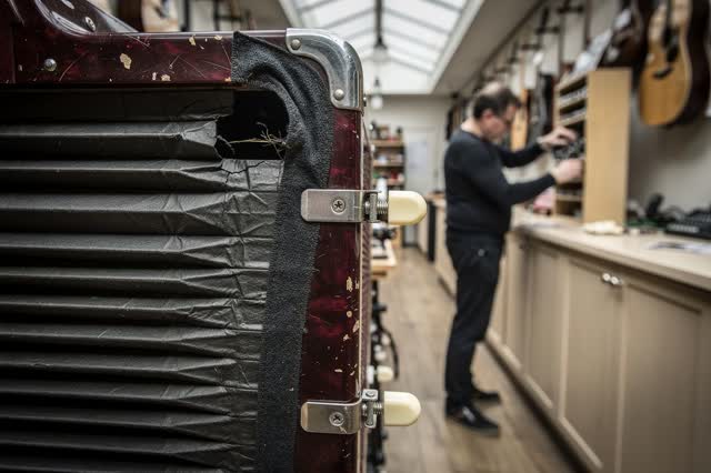 A tender practical repair in a narrow urban music shop under soft dry skylight. Extreme close foreground shows one crack