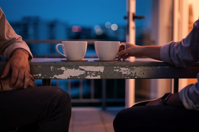 A genuinely quiet image on a modest apartment balcony at blue hour. Two clean white ceramic cups cool on a scuffed metal