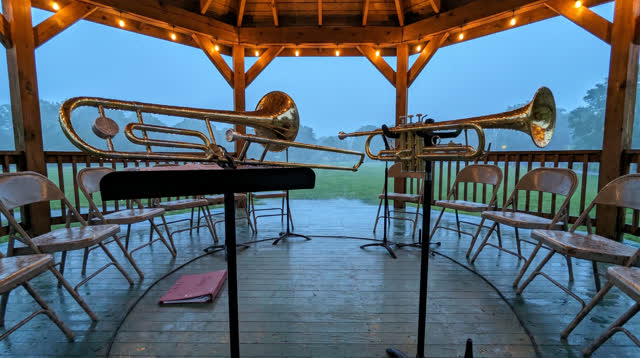 A buoyant first-person dream in an outdoor bandstand during light evening rain. Several used brass instruments rest on s
