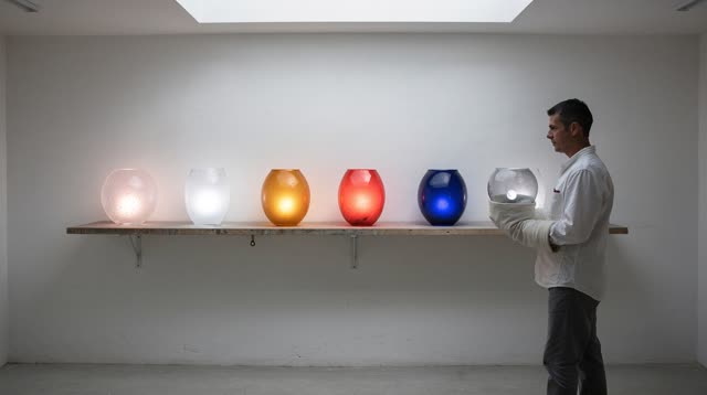 A buoyant first-person dream in a white glassblower's cooling room. Seven clear handblown vessels stand securely along o