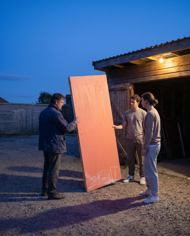 A restrained low-tension challenge in an open rehearsal yard at cool dusk. A lightweight coral-painted prop door stands