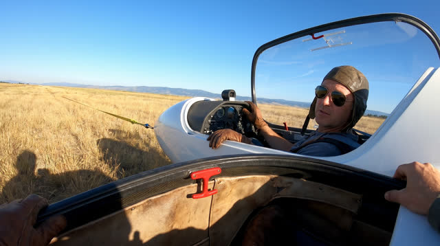 A lucid first-person self-portrait at a high grass airfield in clear morning weather. One sailplane pilot sits securely
