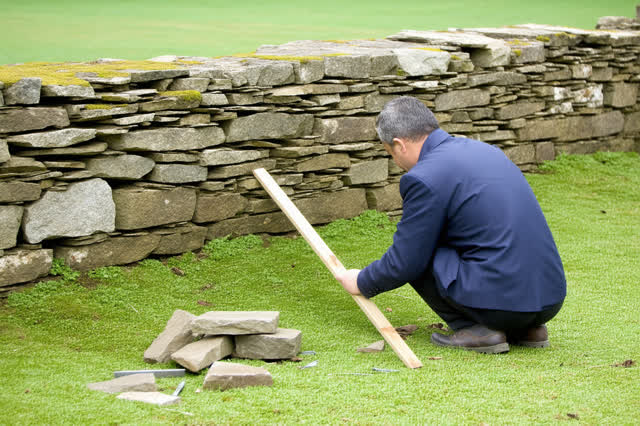 A tender practical repair beside a rural lane in flat bright overcast daylight. A dry-stone mason kneels at one short op