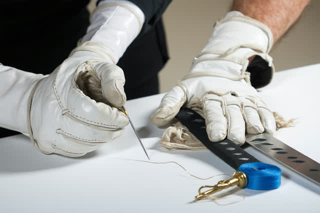 A tender practical repair beside an active fencing competition strip. A coach uses a small curved needle and waxed threa