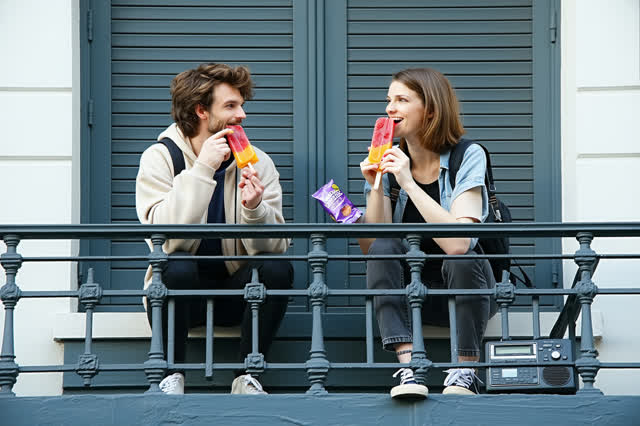 A genuinely quiet summer blue-hour scene on a broad, safe apartment fire escape. Two adult friends sit naturally side by