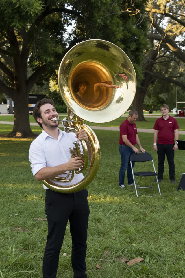 A bright playful scene in a grassy park before a small brass-band rehearsal. One adult musician holds a polished sousaph